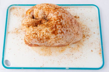 Raw, seasoned turkey breast on a white cutting board with a teal border, sprinkled with various spices and seasoning.
