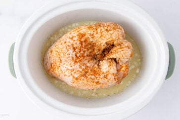 A seasoned raw turkey breast sits in a white slow cooker with chopped onions and liquid at the bottom, ready to be cooked.