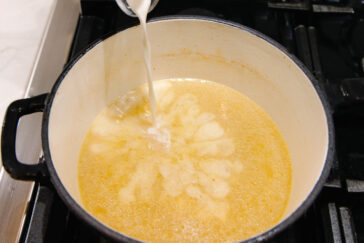 Milk being poured into a pot of bubbling yellow soup or sauce on a stovetop, creating a marbled pattern on the surface.