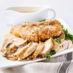 Sliced roasted turkey breast garnished with fresh herbs is served on a white plate with a side of creamy gravy in a white sauce boat in the background. A striped cloth napkin is placed underneath the plate.