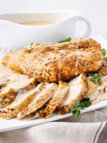 Sliced roasted turkey breast garnished with fresh herbs is served on a white plate with a side of creamy gravy in a white sauce boat in the background. A striped cloth napkin is placed underneath the plate.