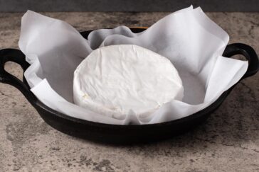A round wheel of brie cheese sits on white parchment paper inside a black cast iron baking dish, placed on a textured gray surface.