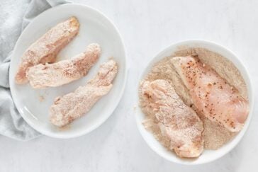 Two white plates on a light surface, one with breaded raw chicken tenders, and the other with raw chicken breasts being coated in breadcrumbs and seasoning. A light gray cloth is partially visible beside the plates.