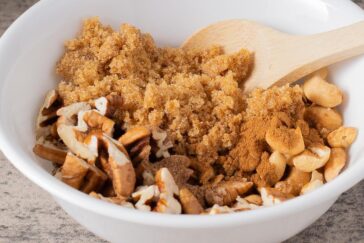 A white bowl contains brown sugar, chopped pecans, peanuts, and ground cinnamon, with a wooden spoon resting inside.