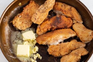 Several pieces of seasoned, breaded chicken are cooking in a skillet with melting butter and minced garlic. The edges of the chicken are golden brown.