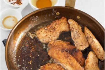 Several pieces of seasoned, browned chicken are frying in a copper skillet with oil. Small bowls with sauce, honey, and chili flakes are visible in the background on a white surface.