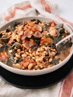 A bowl of black-eyed peas with greens and crispy bacon pieces, served in a light broth, sits on a black plate atop a red and white striped cloth napkin. A spoon rests inside the bowl.