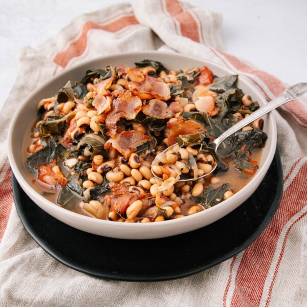 A bowl of black-eyed peas with greens and crispy bacon pieces, served in a light broth, sits on a black plate atop a red and white striped cloth napkin. A spoon rests inside the bowl.