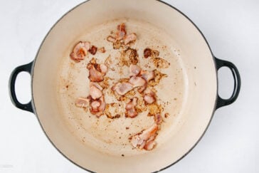 A few pieces of cooked bacon sit in a large, round, cream-colored pot with black handles, showing browned bits and grease residue on the bottom.