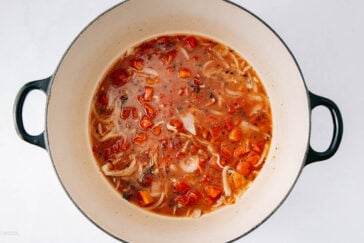 A large pot filled with chunky vegetable soup, featuring visible pieces of tomato, carrot, cabbage, and herbs in a clear broth. The pot sits on a white surface.