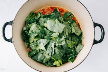 A large pot with chopped collard greens placed on top of a tomato-based mixture, ready to be cooked. The pot has black handles and sits on a white surface.