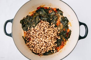 A large pot contains cooked collard greens, sliced onions, diced tomatoes, and a layer of black-eyed peas on top. The ingredients appear to be in the process of being mixed together.