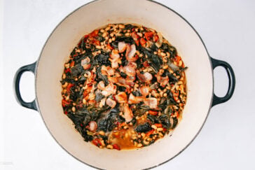 A large pot filled with cooked black-eyed peas, dark leafy greens, diced tomatoes, and pieces of bacon in a savory broth, viewed from above on a white background.