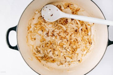 Sliced onions being sautéed with spices in a large cream-colored pot, stirred with a white spoon; the bottom of the pot has browned bits from cooking.