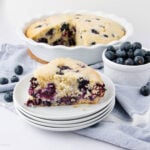 A slice of blueberry cake on a stack of white plates is in the foreground, with the rest of the cake in a pie dish and a small bowl of fresh blueberries in the background. Blueberries are scattered on a light cloth.