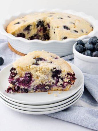 A slice of blueberry cake on a stack of white plates is in the foreground, with the rest of the cake in a pie dish and a small bowl of fresh blueberries in the background. Blueberries are scattered on a light cloth.