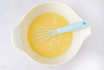 A pale mixing bowl filled with a yellow liquid batter, small lumps visible, and a light blue whisk resting inside. The bowl is placed on a white surface.
