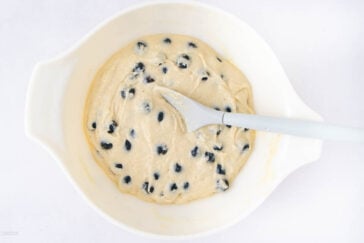 A white mixing bowl with blueberry muffin batter and a white spatula resting in it, set on a white surface. The batter is thick and studded with whole blueberries.