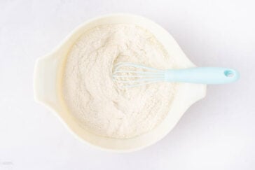 A white mixing bowl filled with flour and a blue-handled whisk resting inside, all placed on a white surface.