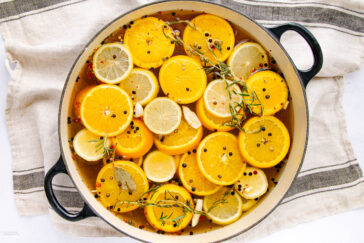 A large pot filled with slices of oranges and lemons, sprigs of rosemary, bay leaves, whole peppercorns, and garlic cloves in liquid, resting on a striped kitchen towel.