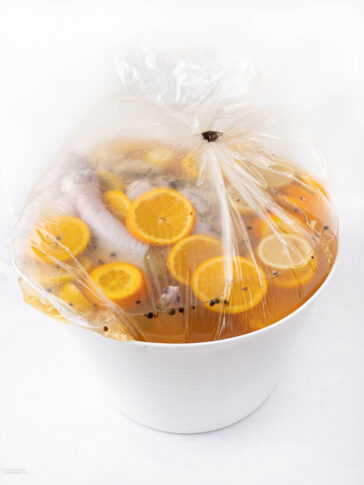 A large white container holds a clear plastic bag filled with liquid, orange and lemon slices, and spices, likely brining poultry or meat. The bag is tightly closed at the top.