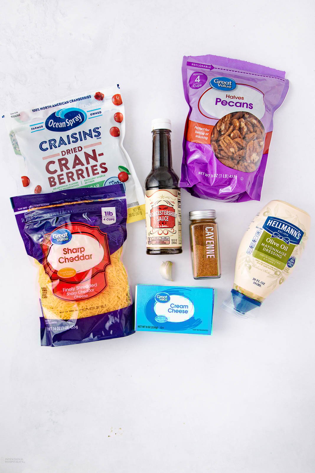 A flat lay of ingredients: a bag of dried cranberries, a bag of pecan halves, shredded sharp cheddar cheese, a bottle of Worcestershire sauce, ground cayenne pepper, a block of cream cheese, and a bottle of mayonnaise.