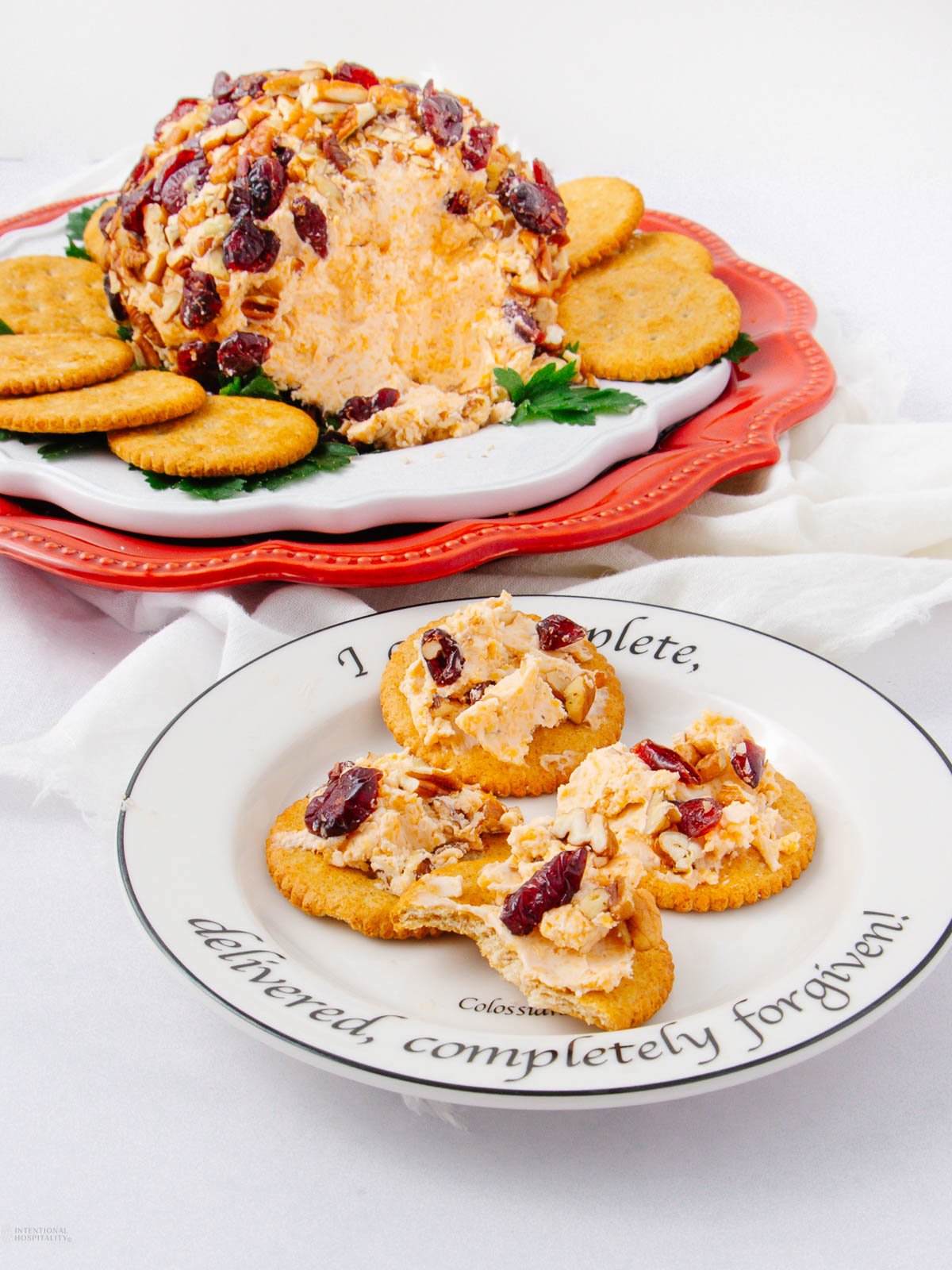 A cheese ball coated with nuts and cranberries, served on a white platter with crackers. In front, three crackers topped with cheese spread are arranged on a decorative plate with a cursive inscription.