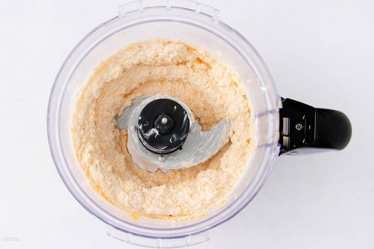 A food processor containing a creamy, light-colored mixture, viewed from above on a white background. The mixture is smooth and partially blended around the blades.