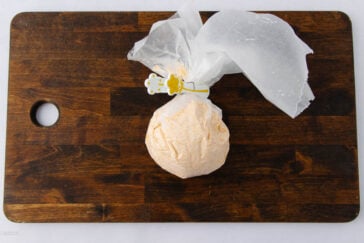 A ball of dough wrapped in parchment paper and secured with a clip sits on a dark wooden cutting board with a circular hole on the left side.