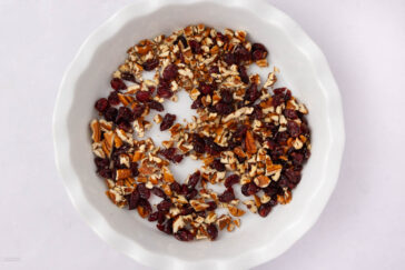 Chopped pecans and dried cranberries are scattered in a white, fluted-edge pie dish on a light background.