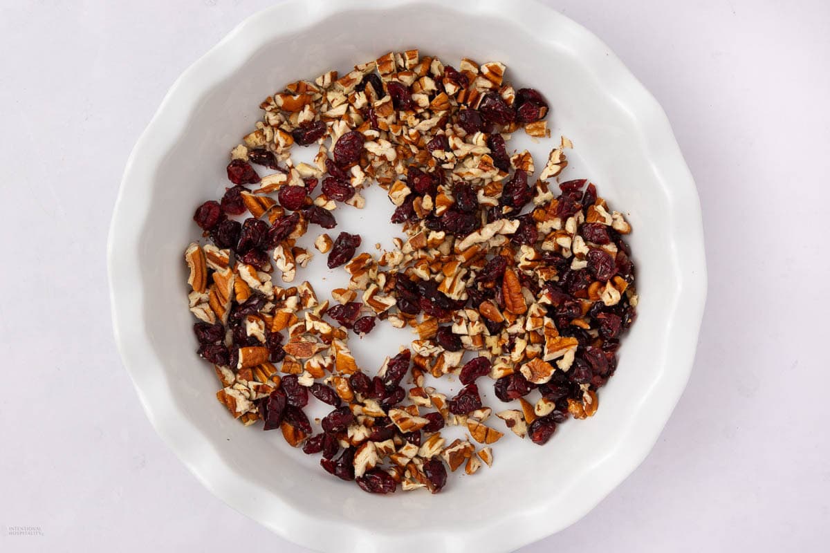 Chopped pecans and dried cranberries are scattered in a white, fluted-edge pie dish on a light background.