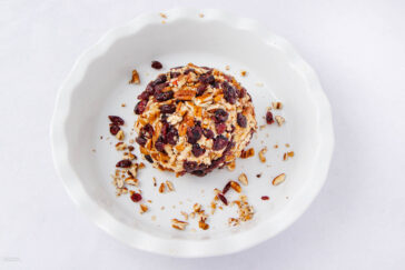 A round cheese ball coated with chopped nuts and dried cranberries sits in a white dish, with some nuts and cranberries scattered around it against a plain white background.
