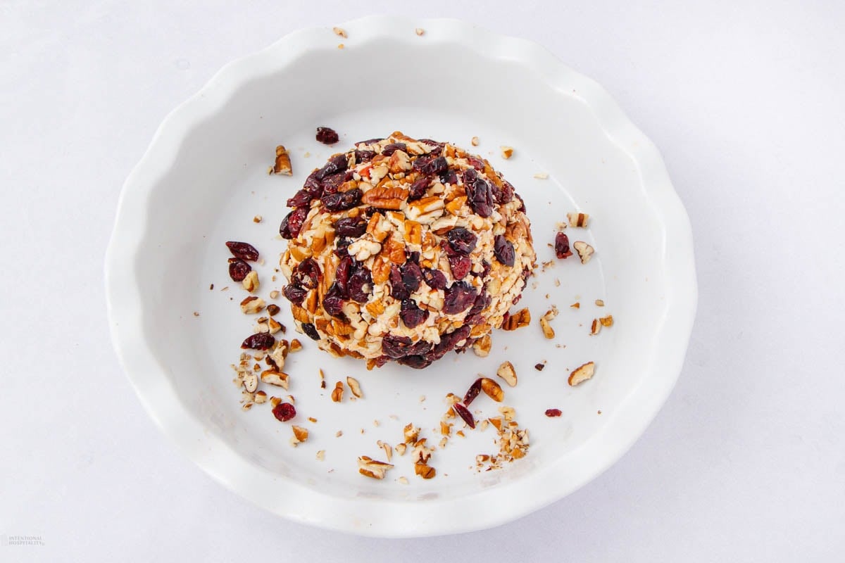 A round cheese ball coated with chopped nuts and dried cranberries sits in a white dish, with some nuts and cranberries scattered around it against a plain white background.