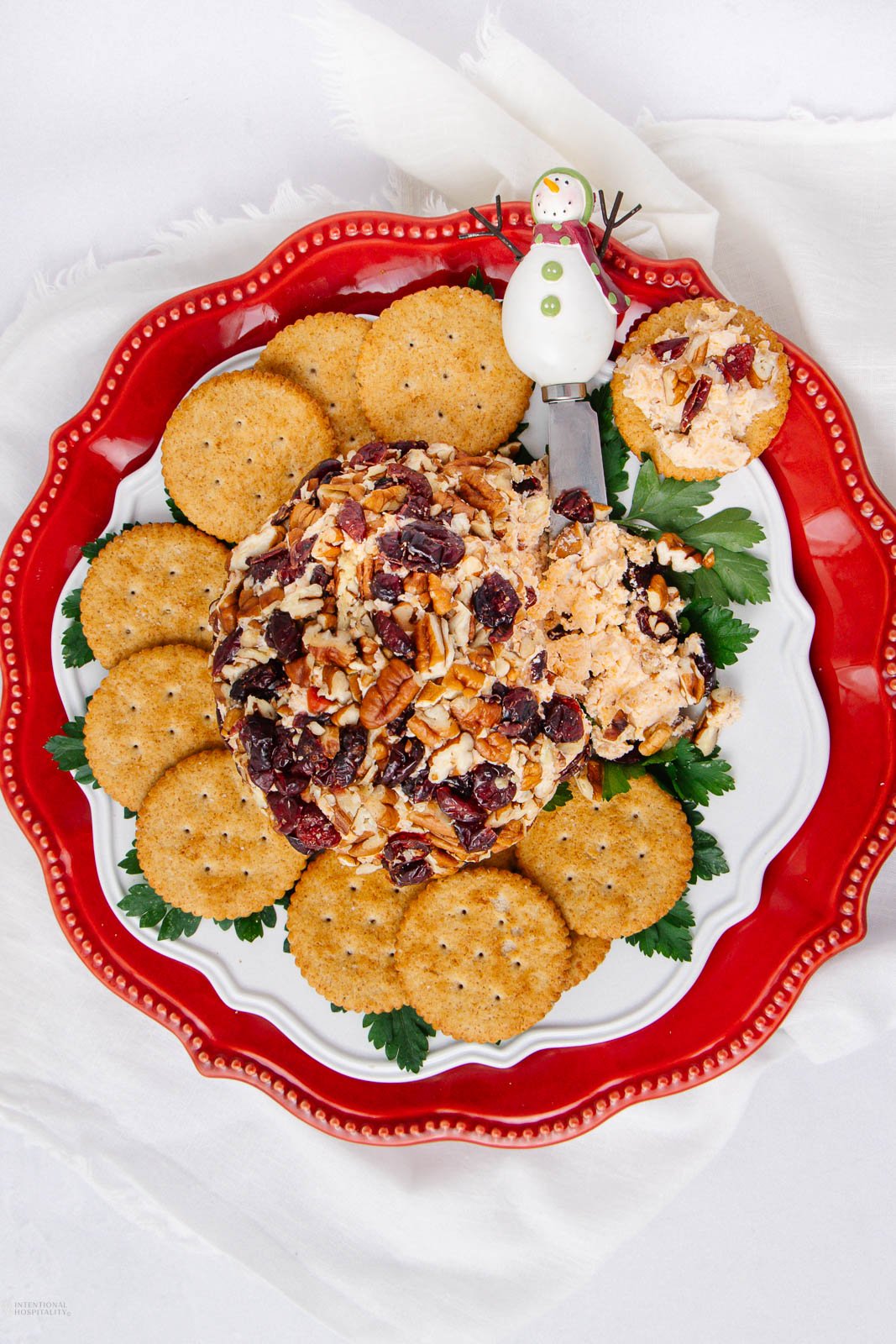 A cheese ball coated with pecans and dried cranberries is surrounded by round crackers on a red and white plate. A snowman-topped spreader knife rests nearby on a white cloth.