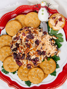 A festive cheese ball covered in nuts and dried cranberries, surrounded by round crackers on a red and white plate, with a small snowman cheese spreader on the side. Green parsley is used as garnish.