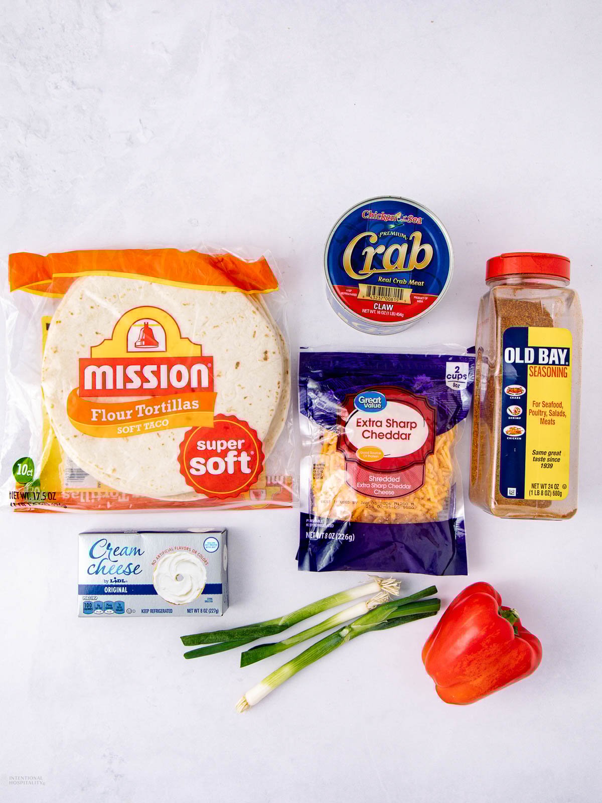 A flat lay of ingredients on a white surface, including flour tortillas, canned crab meat, Old Bay seasoning, cream cheese, shredded cheddar cheese, green onions, and a red bell pepper.