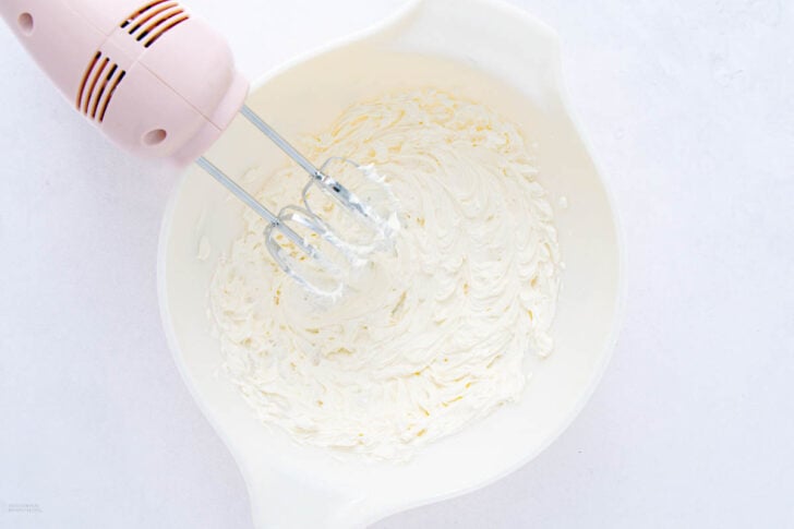 A pink hand mixer is whipping cream in a white mixing bowl on a white surface. The cream appears thick and well-mixed.