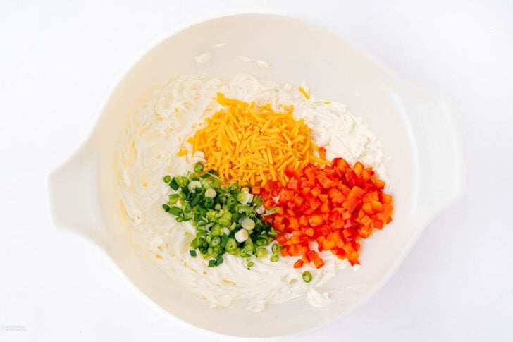 A white mixing bowl with cream cheese spread around the edges, shredded cheddar cheese, chopped green onions, and diced red bell peppers arranged in separate piles.