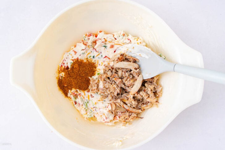 A white mixing bowl containing a creamy mixture with herbs, shredded crab meat, and a pile of reddish-brown seasoning, being stirred with a white spatula.