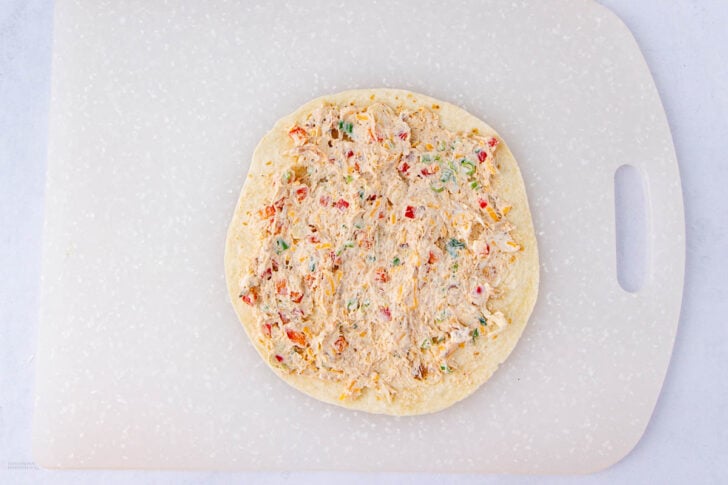 A tortilla spread with a creamy mixture containing shredded chicken and diced colorful vegetables, placed on a white cutting board.