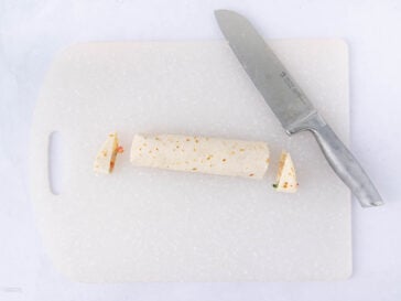 A rolled tortilla wrap with its ends sliced off sits on a white cutting board next to a large kitchen knife.