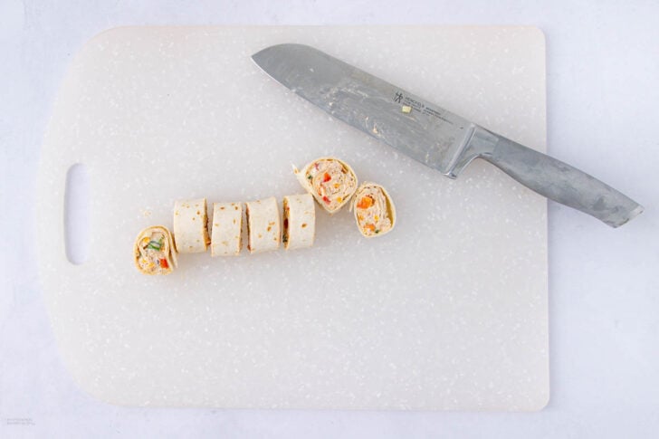 A large chef’s knife rests on a white cutting board next to a sliced tortilla roll-up filled with a colorful mixture, cut into several bite-sized pieces.
