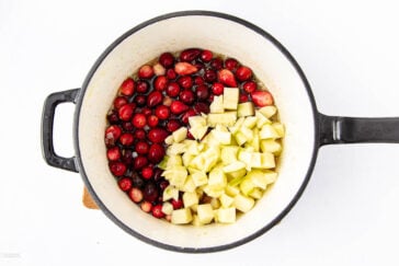 A top-down view of a pot filled half with fresh cranberries and half with chopped green apples, ready to be cooked, on a white background.