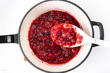 A white saucepan filled with homemade cranberry sauce, featuring whole and crushed cranberries, with a white spoon lifting a portion of the sauce. The background is white.