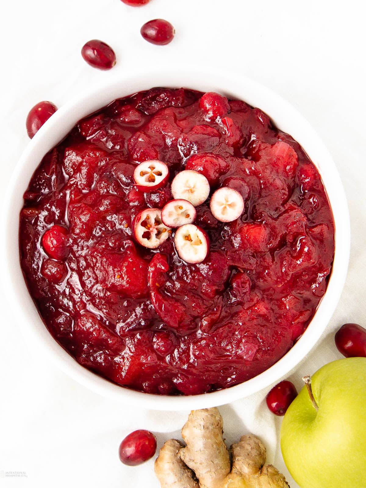 A white bowl filled with chunky cranberry sauce, garnished with halved cranberries. Whole cranberries, a piece of ginger, and part of a green apple are arranged around the bowl on a white surface.