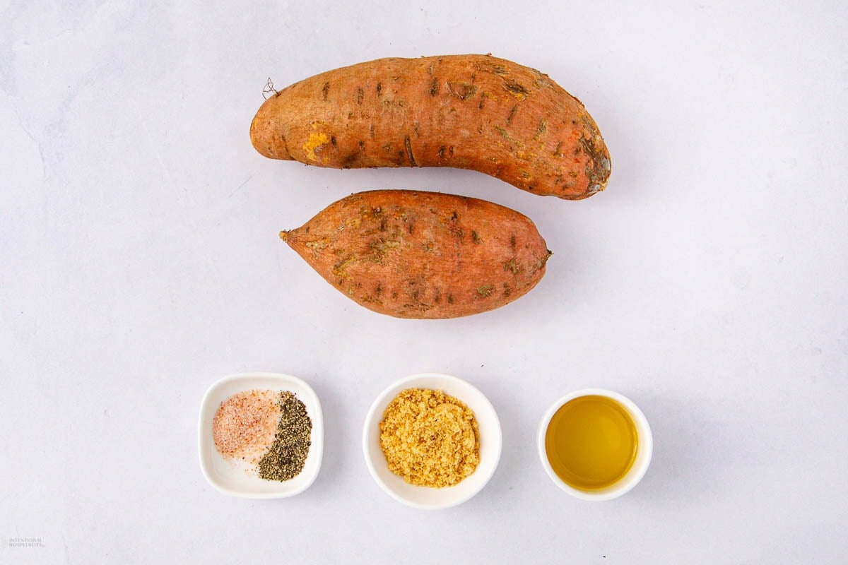 Two whole sweet potatoes placed above three small bowls containing mixed spices, grated cheese, and olive oil, all arranged neatly on a white surface.