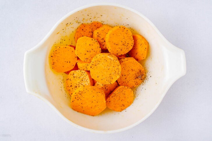 A white bowl filled with sliced sweet potatoes sprinkled with black pepper and seasoning, set on a light background.