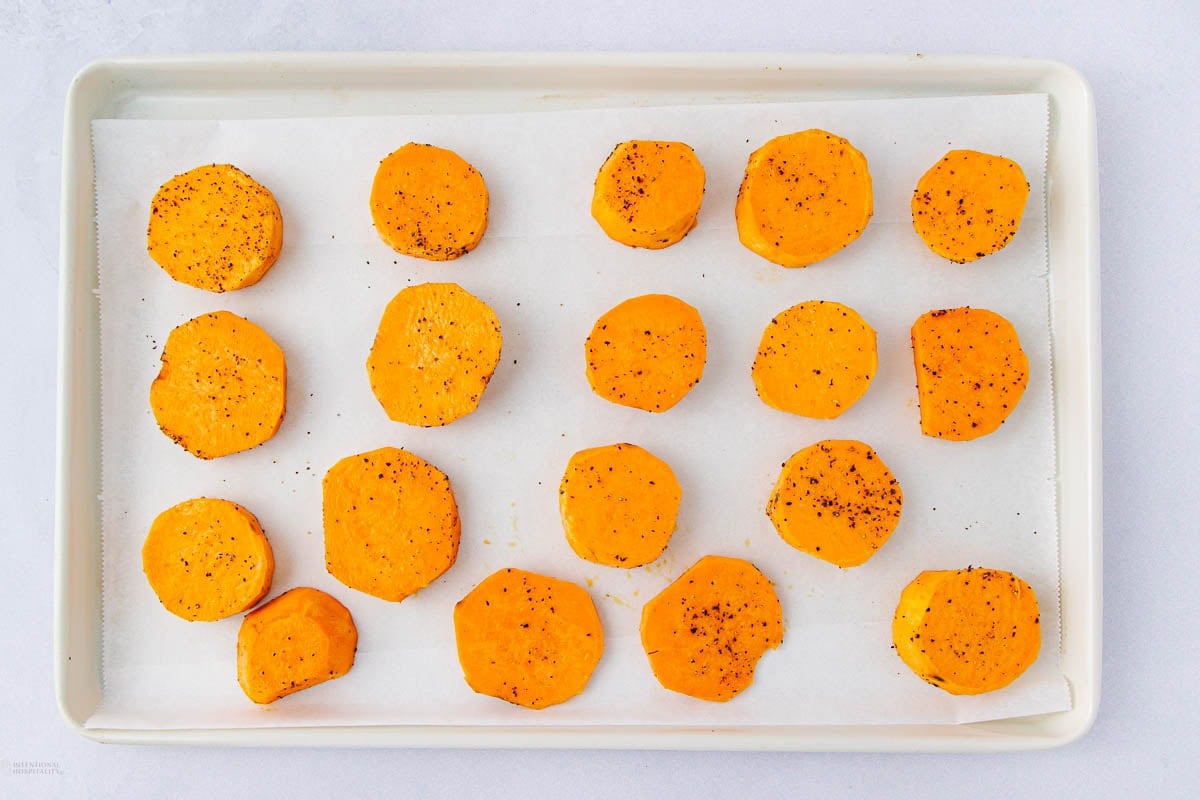 Slices of raw sweet potato arranged in rows on a parchment-lined baking sheet, seasoned with black pepper and spices, ready for roasting.