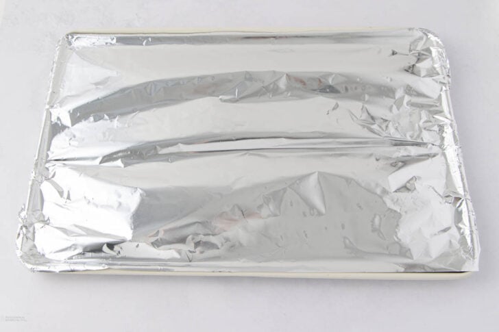 A baking sheet covered tightly with a layer of crinkled aluminum foil, resting on a white surface.