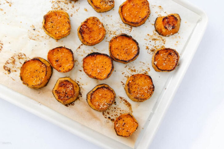 Sliced roasted sweet potatoes with seasoning arranged on a parchment-lined baking sheet. The edges are caramelized, and some slices have a golden-brown crust. The image is bright and taken from above.